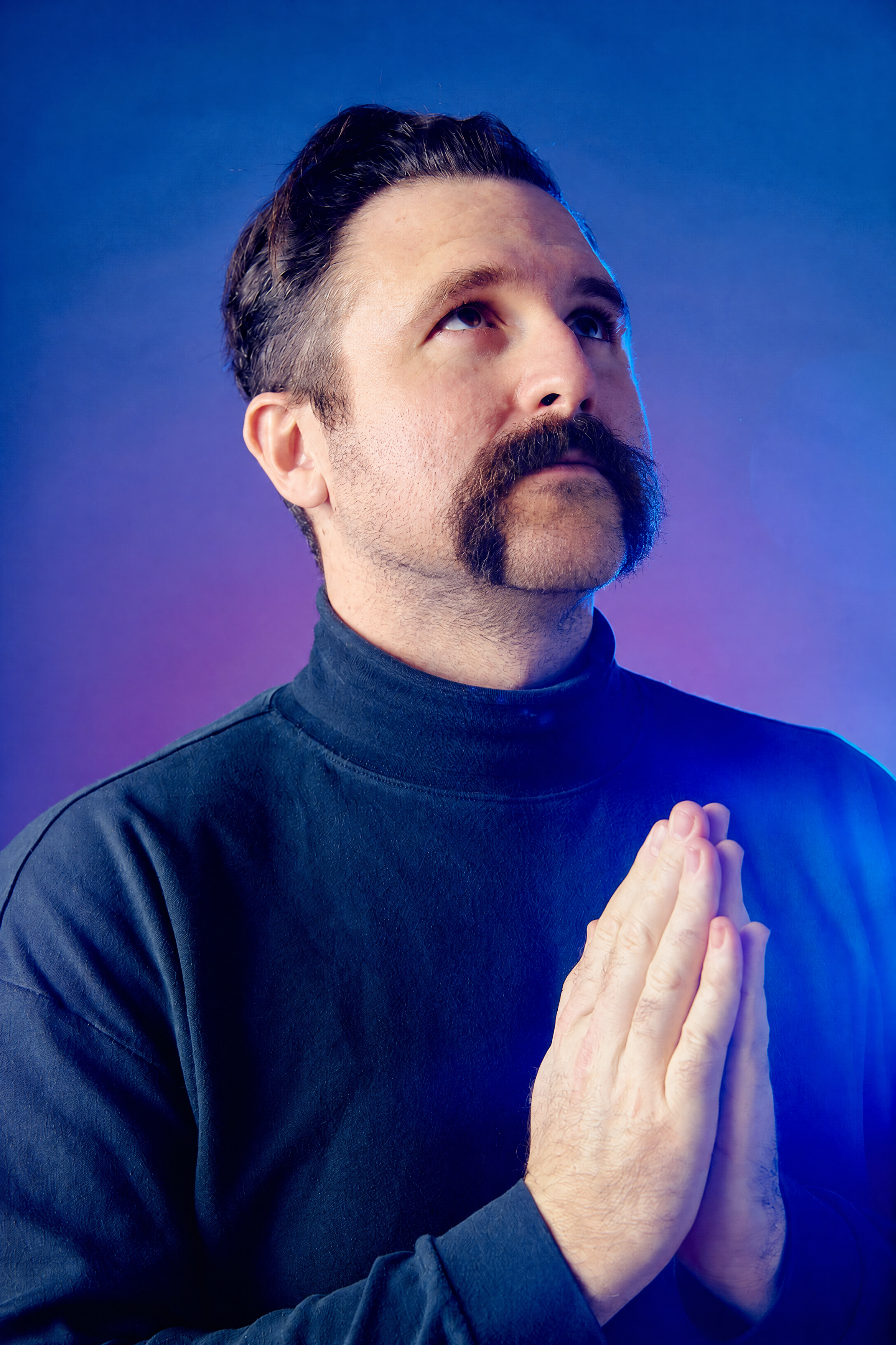 Stuart Bury in blue and purple light, looking upward with hands together
